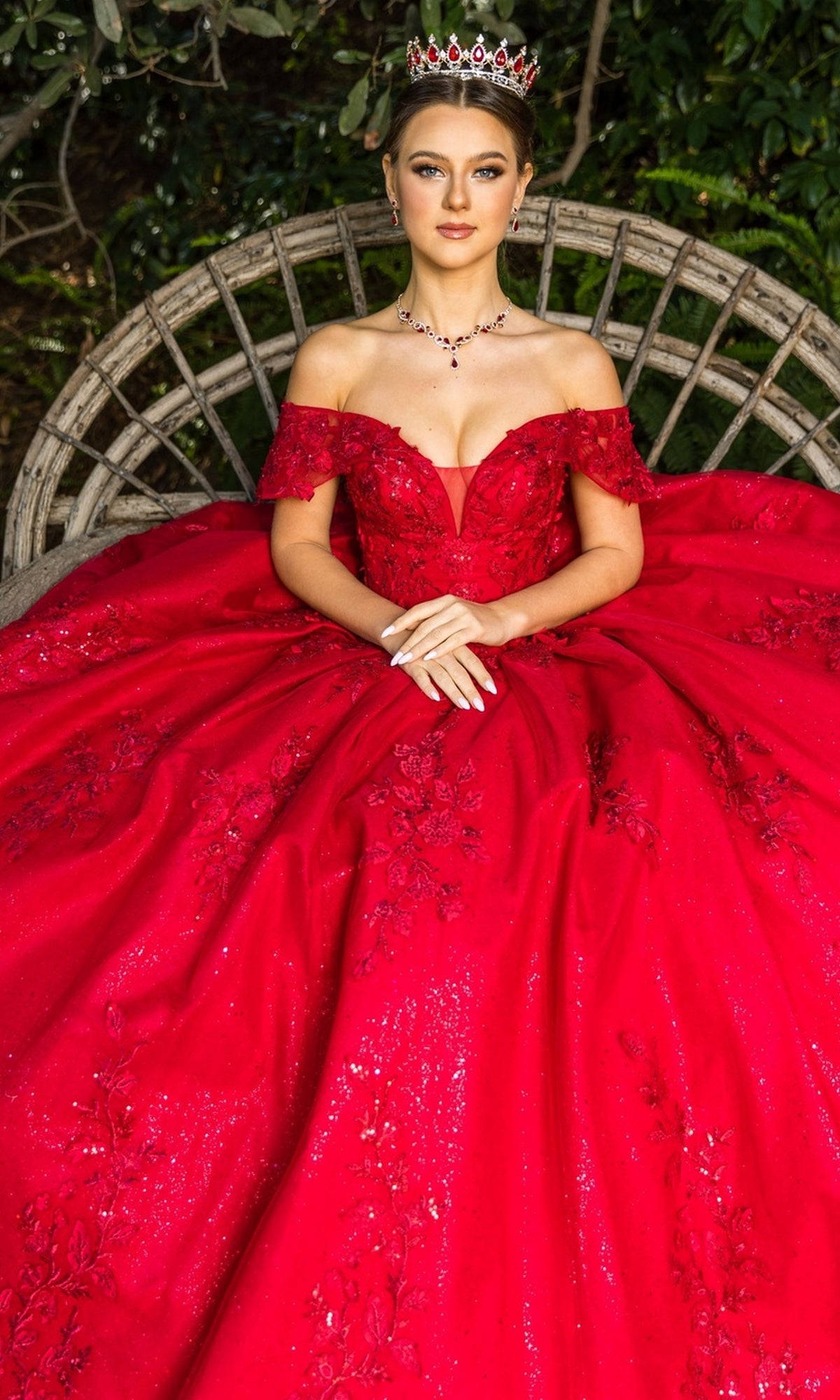 A woman in the Quinceanera Ball Gown 8045J, featuring red off-the-shoulder glitter lace embroidery and a jeweled crown, sits on a round wooden bench with greenery behind her.